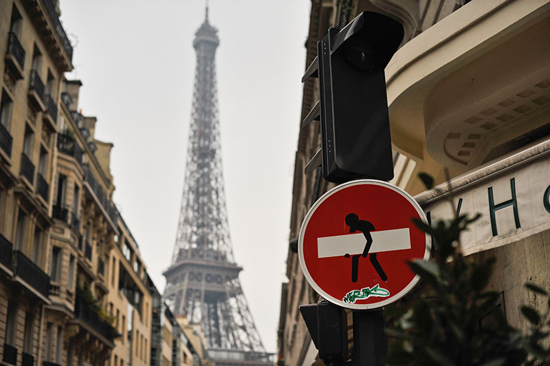 Unusual view of the Eiffel Tower, with man carrying a bar on a street sign. Local graffiti